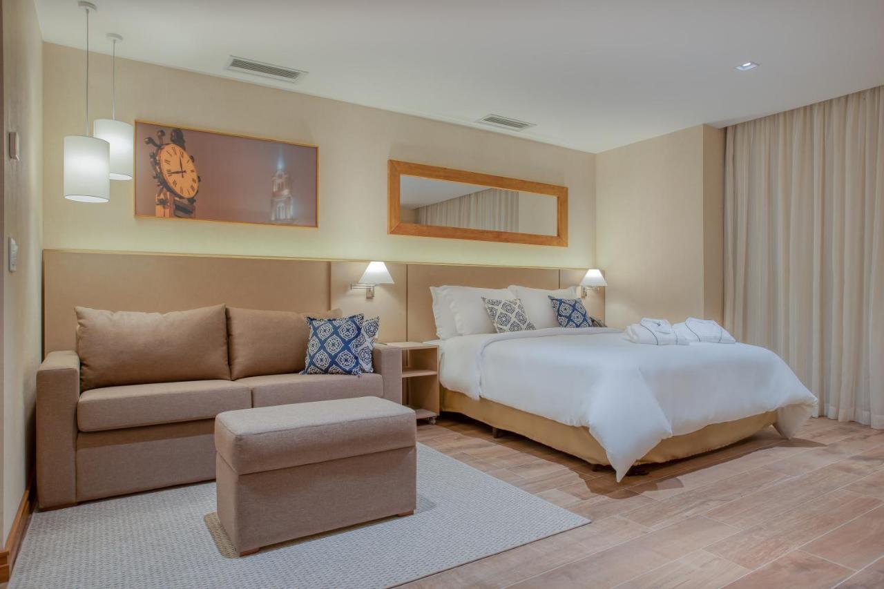 Wyndham Gramado Termas Resort & Spa Ngoại thất bức ảnh The photo shows a modern hotel room featuring a cozy, well-decorated interior. There’s a large bed with white bedding, complemented by decorative pillows. Next to the bed is a stylish nightstand with a lamp. A comfortable sofa is positioned in one co