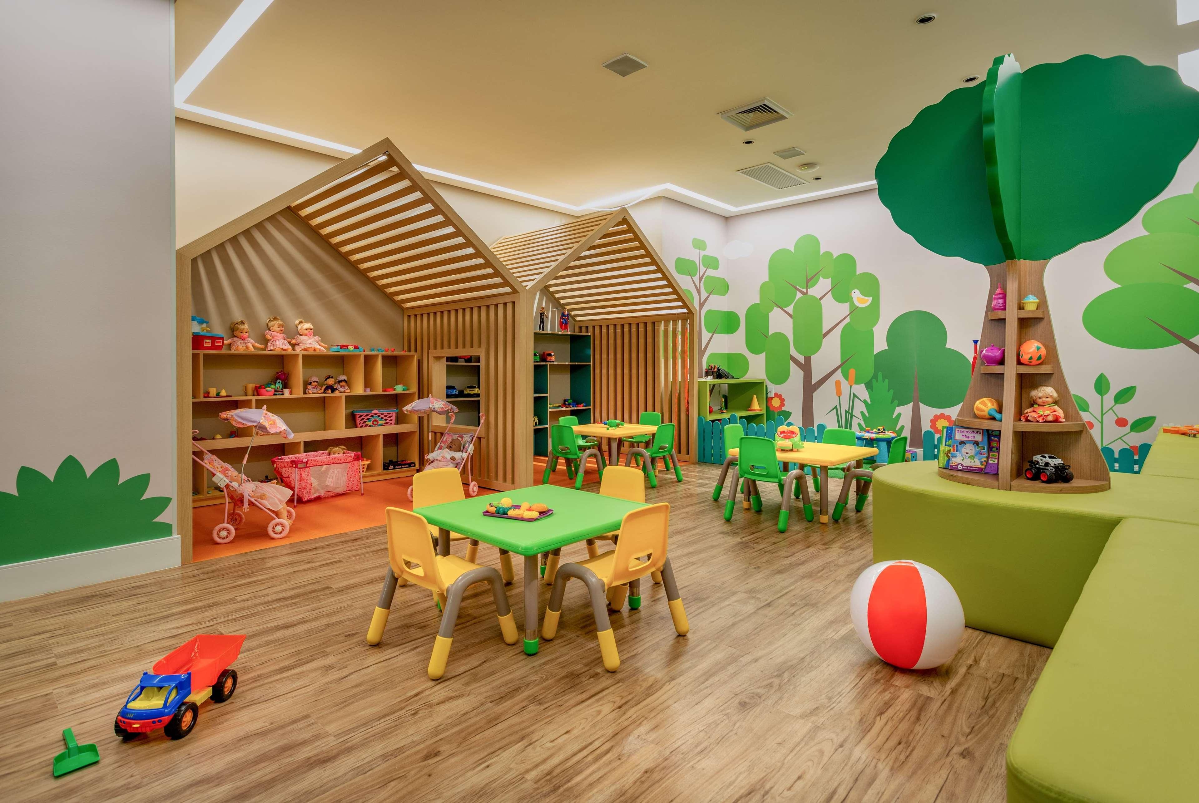 Wyndham Gramado Termas Resort & Spa Ngoại thất bức ảnh The photo shows a colorful playroom designed for children. It features two wooden playhouses with a slatted roof, a shelving unit filled with toys, and several small tables with chairs. The tables are surrounded by bright green chairs, and there are 
