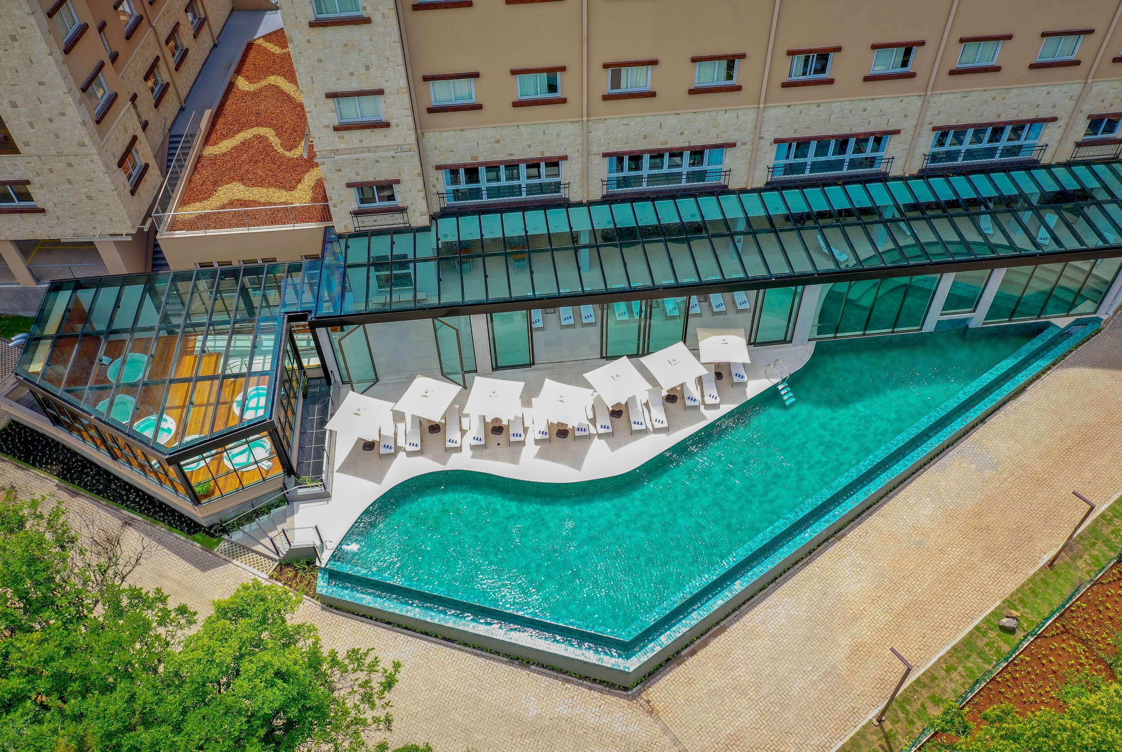 Wyndham Gramado Termas Resort & Spa Ngoại thất bức ảnh The image shows a modern indoor swimming pool area located alongside a building. The pool has an elegant, curvy shape and is surrounded by several white lounge chairs. Large glass panels provide a view of the pool, allowing natural light to flood the