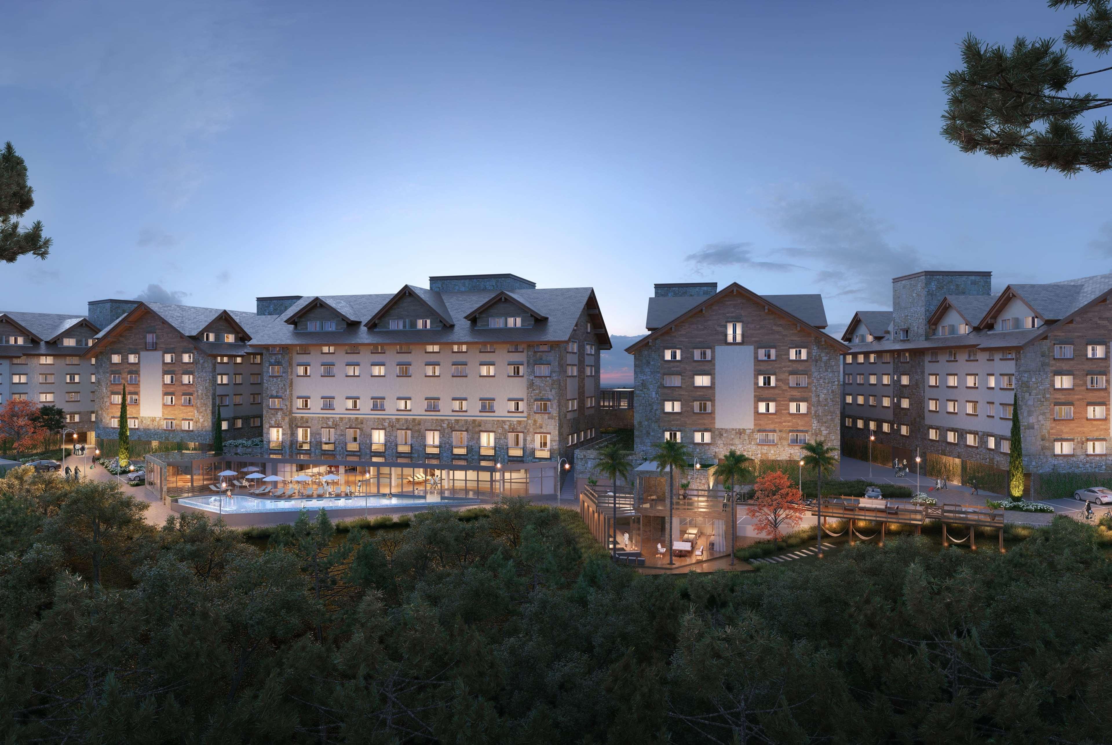 Wyndham Gramado Termas Resort & Spa Ngoại thất bức ảnh The photo depicts a modern hotel complex nestled in a natural setting. The buildings have a contemporary design with a mix of stone and wooden elements, featuring sloped roofs. It appears to be dusk, as the sky has a soft glow, enhancing the aestheti