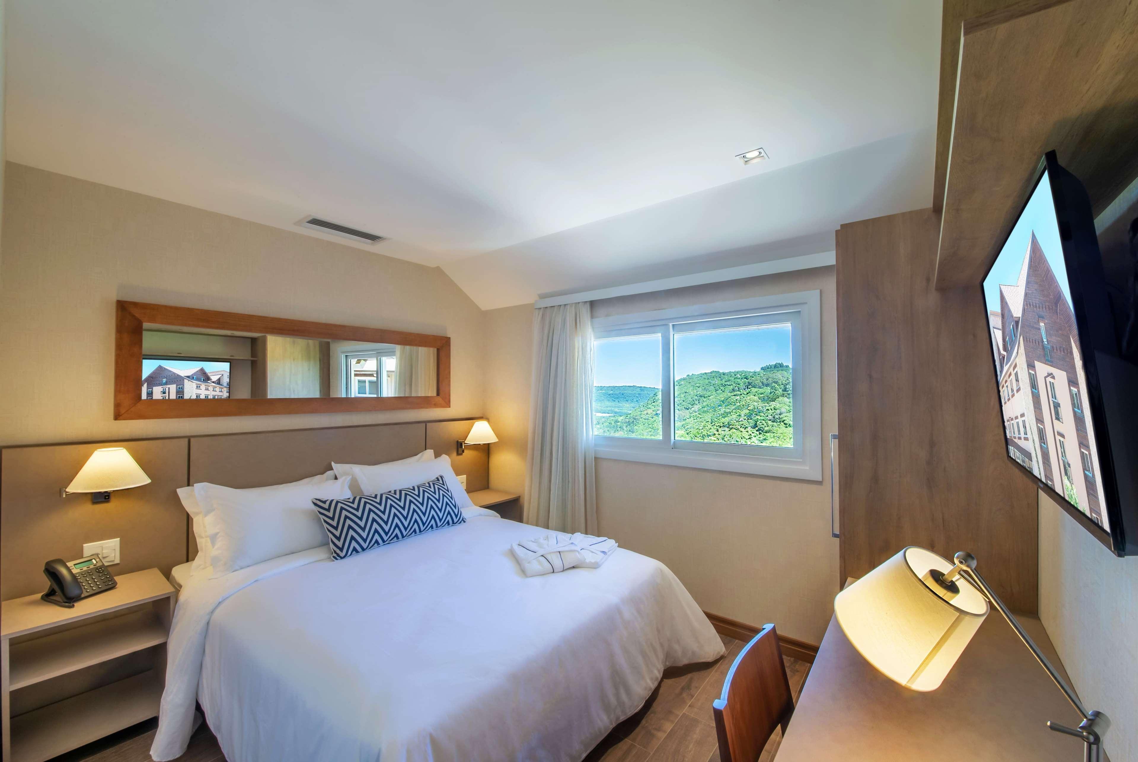 Wyndham Gramado Termas Resort & Spa Ngoại thất bức ảnh The photo shows a cozy hotel room featuring a queen-size bed with white linens and a decorative pillow. On either side of the bed are bedside lamps. There is a desk with a modern desk lamp and a flat-screen television mounted on the wall. A window le