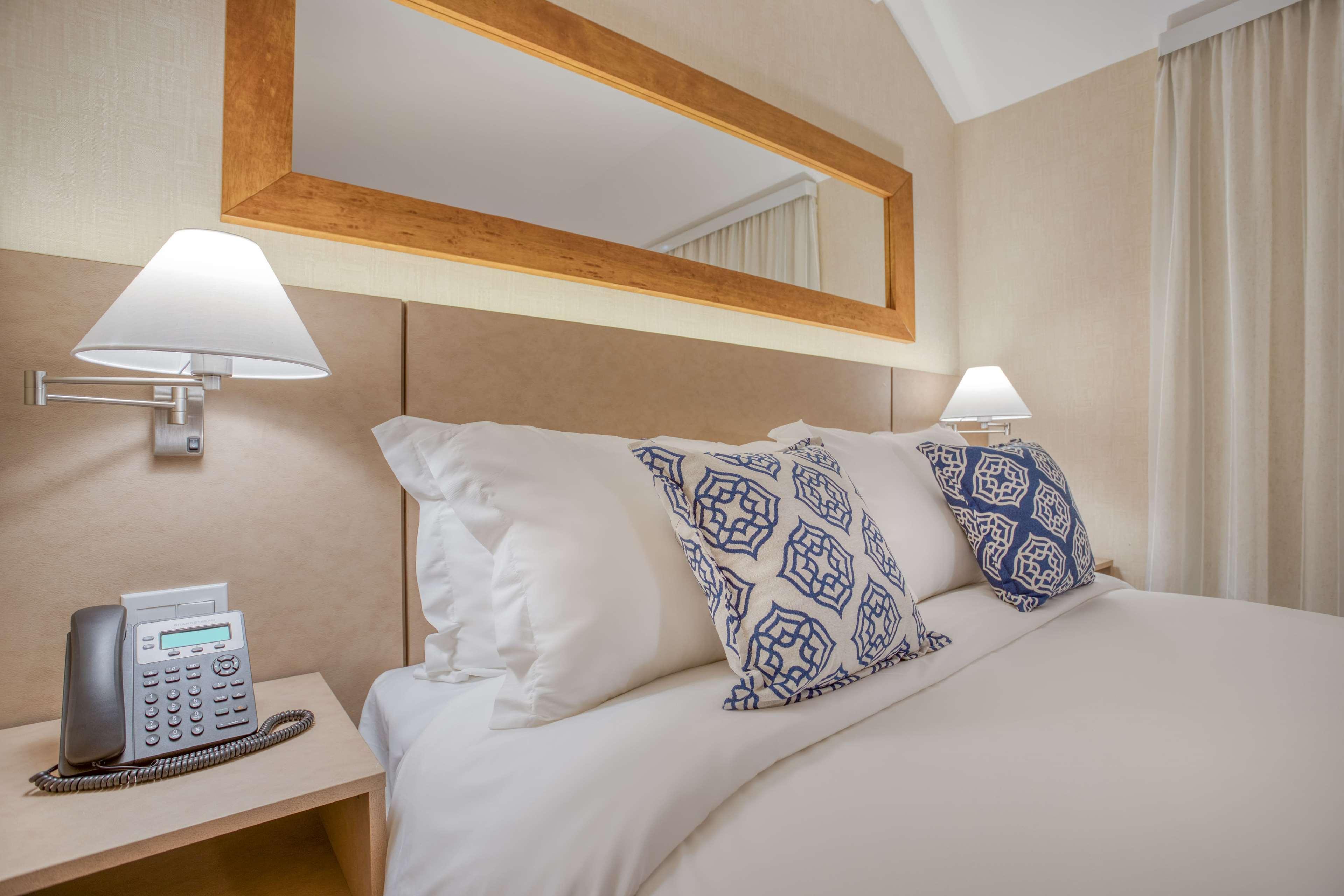 Wyndham Gramado Termas Resort & Spa Ngoại thất bức ảnh The photo shows a cozy hotel room with a neatly made bed. The bed is adorned with large pillows featuring blue geometric patterns. There are bedside lamps on either side, providing soft lighting. A wooden mirror is mounted on the wall above the bed, 