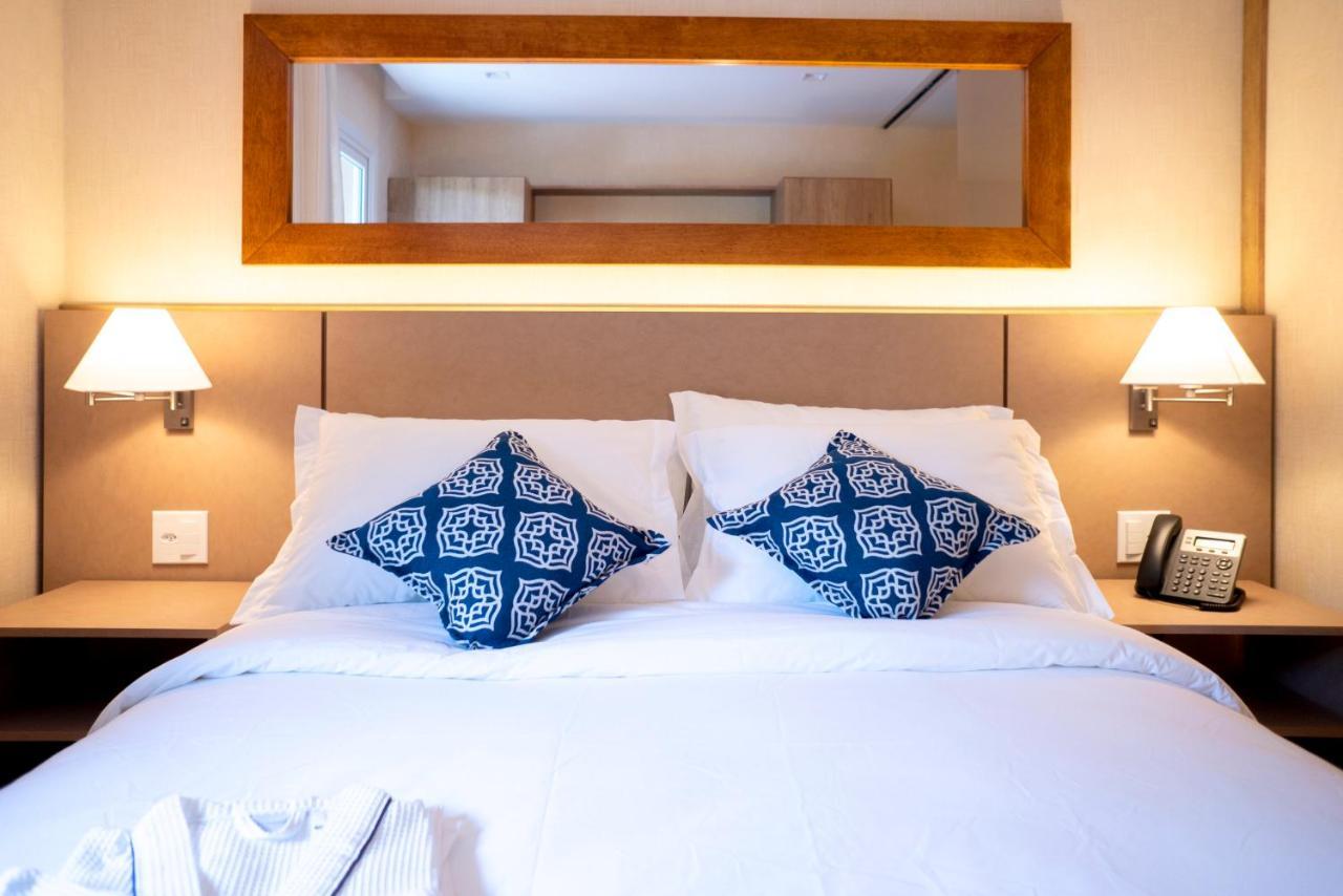Wyndham Gramado Termas Resort & Spa Ngoại thất bức ảnh The photo shows a neatly made bed with white linens and two decorative pillows featuring a blue geometric pattern. Above the bed, there is a wooden-framed mirror reflecting the room's interior. On either side of the bed, there are wall-mounted lamps 