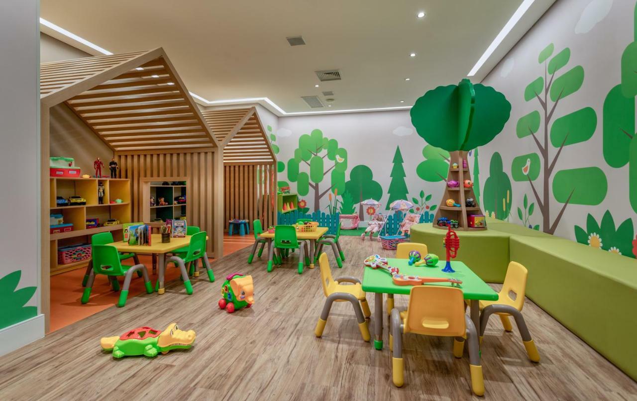 Wyndham Gramado Termas Resort & Spa Ngoại thất bức ảnh The photo shows a colorful children's playroom. It features several small tables and chairs in green and yellow, arranged for play. The walls are adorned with bright green tree and plant designs, giving the room a nature-themed atmosphere. Play mater